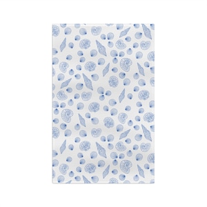 May include: White rectangular fabric with a repeating pattern of blue seashells. The design includes various shell shapes, such as spiral, fan, and cone, scattered across the surface. The shells are outlined in blue.
