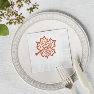 May include: A white paper napkin with a leaf design in orange, featuring the words "FUCK TRUMP". The napkin is placed on a white plate with a gold rim, alongside a fork and knife.