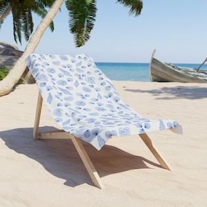 May include: A white beach chair with a blue and white seashell pattern. The chair is on a sandy beach with palm trees and a blue ocean in the background.