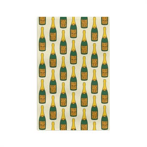 May include: A rectangular, cream-colored decorative towel featuring a repeating pattern of champagne bottles. Each bottle has a green body and a gold neck, with text that reads "IS HE DEAD YET?" and "FUCK TRUMP."
