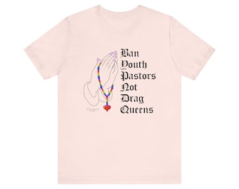 May include: Pink t-shirt featuring a graphic of praying hands holding a rainbow rosary with a red heart charm. The text "Ban Youth Pastors Not Drag Queens" is printed in black.