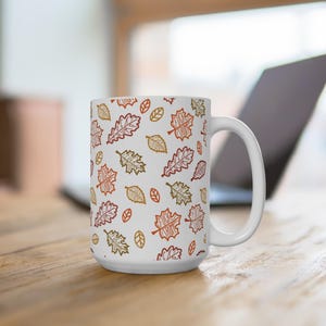 Pode incluir: Caneca de cerâmica branca com um padrão repetido de folhas de outono em tons de laranja, vermelho e dourado. As folhas são estilizadas e espalhadas pela superfície da caneca. A caneca tem uma alça grande e curva.