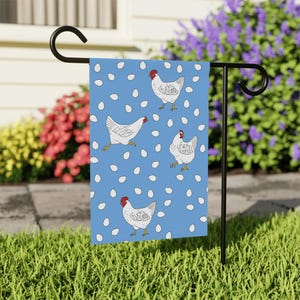 May include: A blue garden flag featuring a repeating pattern of white chickens and white eggs. Each chicken has a red comb and the text "fuck around" printed on its body. The flag is displayed on a black metal stand in a garden setting.