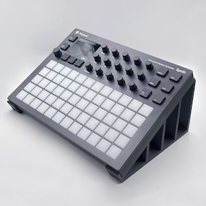 May include: A black Roland Boutique Synthesizer with a white grid of keys and a small screen. The synthesizer is on a black stand with three legs.