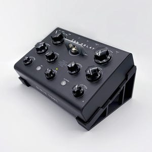 Può includere: Un pedale per effetti audio nero con più manopole e la scritta "ZEN DELAY" in alto. Il pedale ha una forma rettangolare con una serie di manopole e interruttori. Il dispositivo è progettato per strumenti musicali.
