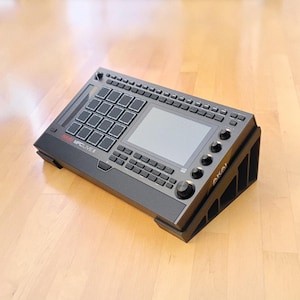 May include: A black Akai MPC Live II music production center with a screen, pads, and knobs. The device is on a black stand, angled slightly. The text "MPC LIVE II" is visible. The device is on a wooden surface.