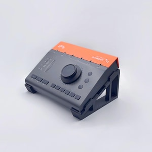 May include: A gray and orange audio interface with a large central knob and several buttons. The device is angled and supported by black stands. The top section is orange, and the device has the text "Studio Connect 72".