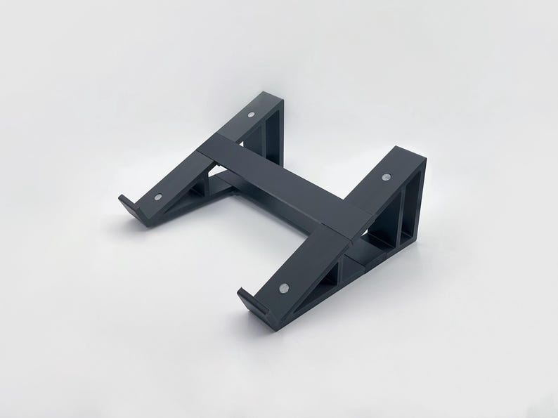 May include: A dark gray laptop stand with an H-shaped design. The stand is made of a solid material and features angled supports with small, silver-colored fasteners. The stand is designed to elevate a laptop for improved ergonomics.
