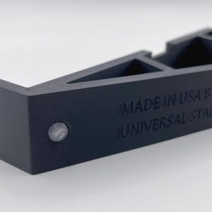 May include: A dark navy blue universal stand with a geometric design. The text "MADE IN USA BY PINE 3D" and "UNIVERSAL STAND - RIGHT" is visible. A small, silver-colored circular element is on the side.