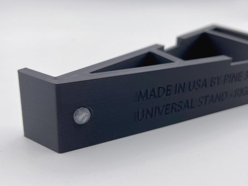 May include: A dark navy blue universal stand with a geometric design. The text "MADE IN USA BY PINE 3D" and "UNIVERSAL STAND - RIGHT" is visible. A small, silver-colored circular element is on the side.