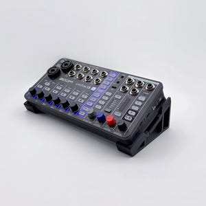 Puede incluir: Una mesa de mezclas digital Zoom LiveTrak L8 negra con múltiples conectores de entrada, perillas y faders. La mesa de mezclas tiene un soporte incorporado y presenta el texto "Zoom LiveTrak L8". La mesa de mezclas tiene perillas azules, rojas y grises.