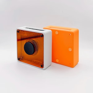 May include: A square device with a white frame and a translucent amber front panel featuring a black circular knob in the center. Beside it stands a solid orange square block with three circular indentations on its side surface.