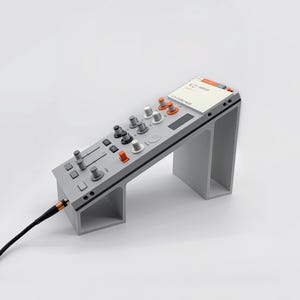 May include: A gray electronic audio mixer with orange and white knobs and sliders. The mixer has a screen displaying text and is connected to a black cable with a gold-colored connector. The mixer is angled and supported by a gray stand.