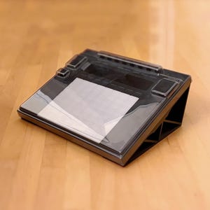 以下が含まれることがあります： 透明なプラスチックカバーと方眼紙のインサートが付いた黒い描画タブレット。 タブレットには内蔵スタンドがあり、上向きに角度が付けられています。 タブレットは木製の表面にあります。