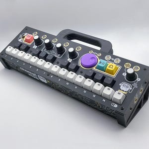 Puede incluir: Un dispositivo de producción musical blanco y negro con una perilla morada, botones amarillos y un teclado blanco etiquetado del 1 al 14. El dispositivo tiene un asa y el texto "Chromatic Club" impreso en la parte delantera.