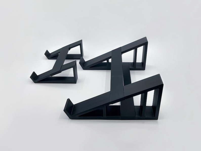May include: Three black geometric display stands of varying sizes are arranged on a white surface. Each stand has a sloped top and a supportive base, creating an angled design. The stands are made of a solid, matte black material.