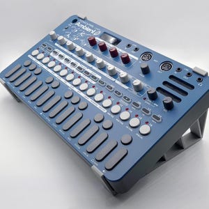 Może przedstawiać: Niebieski syntezator z podstawką, wyposażony w wiele pokręteł, przycisków i suwaków. Syntezator ma wyświetlacz cyfrowy i napis "Ambient 1" nadrukowany z przodu.