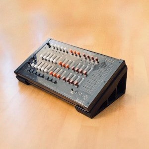 May include: A black audio mixing console with multiple sliders, knobs, and buttons. The console has a rectangular shape and is set on a black stand. The sliders have red and white accents. The console is on a light brown wooden surface.