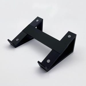 May include: A black, angled stand with a horizontal bar connecting two upright supports. The stand has a minimalist design and appears to be made of a matte material. Small, silver-colored circular accents are visible on the stand.
