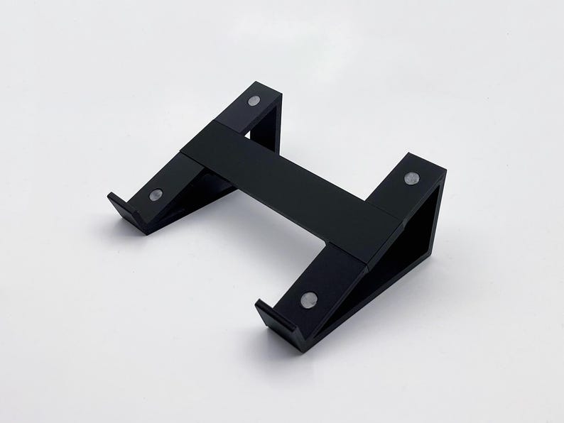 May include: A black, angled stand with a horizontal bar connecting two upright supports. The stand has a minimalist design and appears to be made of a matte material. Small, silver-colored circular accents are visible on the stand.