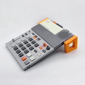 May include: A gray electronic music sampler with black and orange buttons, and the text "KO II" on a white card. The device is supported by an orange stand. The sampler has a grid of black buttons and several control knobs.