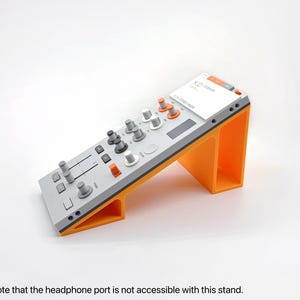 May include: A gray and orange DJ controller stand. The stand is angled and features a gray control surface with knobs and sliders. The stand itself is bright orange. A white label with text is visible on the top.