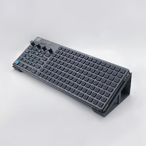 May include: A black keyboard with a grid of keys and a row of knobs. The keyboard is angled on a black stand with the word "ONE" printed on the side.