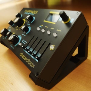 Puede incluir: Sintetizador negro y azul con el texto "TYPHON" y "Dreadbox" en la parte delantera. El sintetizador tiene perillas, botones y deslizadores. Est&aacute; sentado en un soporte negro.