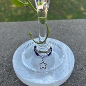 May include: A circular wine glass charm featuring a silver-toned star pendant. The hoop is decorated with a sequence of small beads in shades of dark blue, light purple, and black, accented with tiny silver-colored metal spacers.