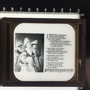 Puede incluir: Una diapositiva de vidrio rectangular con una ilustración en blanco y negro de cuatro figuras con cofias blancas y vestidos oscuros sosteniendo libros. El texto dice I NEED Thee every hour, Most gracious Lord, e incluye notación musical.