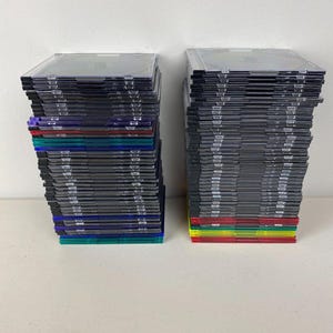 May include: Two stacks of clear plastic CD cases with black inserts. The cases have colored edges, including red, green, blue, and yellow. The cases are stacked on a white surface.