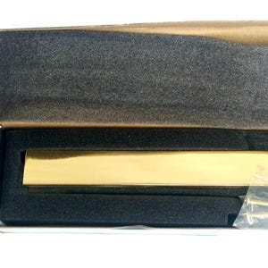 Könnte beinhalten: Ein rechteckiger Briefschlitz aus poliertem, goldfarbenem Metall mit einer glatten, reflektierenden Oberfläche. Der Artikel befindet sich in einem offenen Karton, der mit schwarzem Schaumstoff ausgekleidet ist, inklusive zwei kleiner Schrauben.