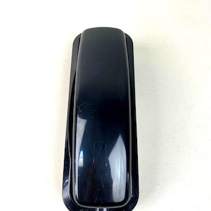 May include: A black, vintage-style telephone handset with a rectangular base. The handset is glossy and reflects light, suggesting a plastic or similar material. The design is simple and classic, suitable for home or office use.