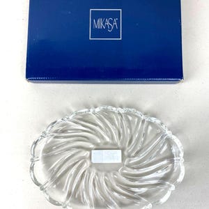 May include: A clear glass serving dish with a swirling design and scalloped edges, next to a dark blue box with the brand name "MIKASA" in a white square.