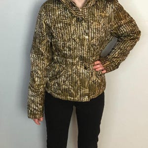 Peut inclure: Une veste à motif doré et noir métallisé dotée d'un col montant avec une bordure en fausse fourrure marron foncé, de manches longues, d'une fermeture éclair frontale avec des boutons métalliques et d'une taille cintrée. Finition texturée.