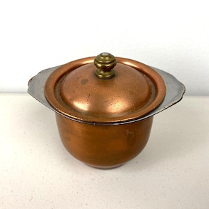Puede incluir: Un pequeño cuenco de color cobre con tapa. La tapa tiene un pomo de latón y un borde plateado. El cuenco es redondo con un borde ligeramente ensanchado. El artículo parece ser vintage o antiguo.