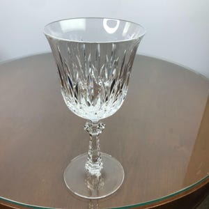May include: A clear crystal wine glass featuring a flared bowl with vertical cut patterns and a faceted stem. The glass has a smooth, circular base and a wide, open rim, showcasing intricate light-reflecting details throughout the body.