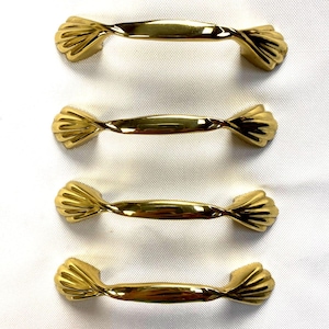 May include: A set of four gold-toned metal cabinet pulls, each featuring a smooth, twisted centre bar and decorative, fan-shaped ends with ribbed detailing. These hardware pieces have a polished, reflective finish and a uniform, elongated shape.