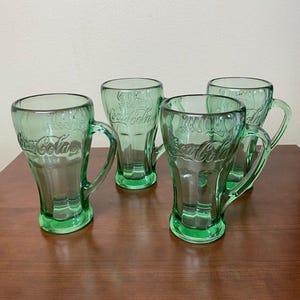 Könnte beinhalten: Vier grüne Coca-Cola-Trinkgläser mit Griffen. Die Gläser bestehen aus durchscheinendem grünen Glas und tragen das Coca-Cola-Logo. Die Gläser stehen auf einer braunen Holzoberfläche.
