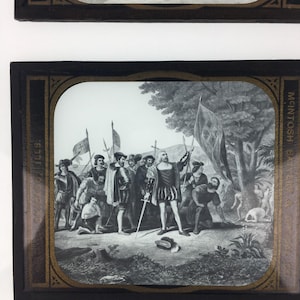 May include: A rectangular glass lantern slide featuring a black and white historical illustration of explorers landing on a shore. The frame is black with gold accents and text reading MCINTOSH BATTERY & OPTICAL CO. CHICAGO ILLS.