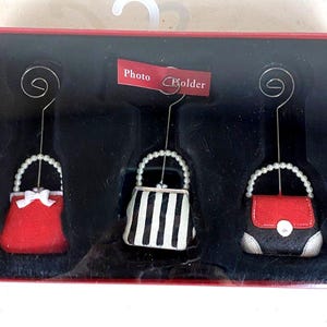 May include: Set of three photo holders shaped like miniature purses. One is red with a white bow, another has black and white stripes, and the third is red and black. Each purse has a pearl handle and a wire top.