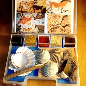 Puede incluir: Una caja de almacenamiento de plástico transparente con un forro de fieltro azul. La caja contiene cuatro pequeños recipientes de polvo de pigmento, cuatro conchas, una pieza de sílex, un pequeño pincel y un trozo de madera. La caja también incluye una hoja de papel con imágenes de pinturas rupestres de Lascaux y la Grotte Chauvet.