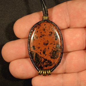 Könnte beinhalten: Ovaler Anhänger mit poliertem, rotbraunem Stein mit schwarzen Einschlüssen. Der Stein ist in einem schwarzen, gewebten Rahmen gefasst und an einer schwarzen Kordel aufgehängt. Messingfarbene Metallakzente.