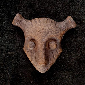 Può includere: Una maschera in metallo marrone, usurata, con un volto di animale stilizzato. La maschera presenta due cavità oculari rotonde, un naso triangolare e due piccole corna rivolte verso l'alto. La superficie ha un aspetto ruvido e strutturato, che suggerisce una qualità invecchiata o antica.