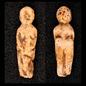 May include: Two small, light-colored figurines of a human form. The figures have rounded heads, arms crossed over the chest, and simplified facial features. The figurines are on a black background.