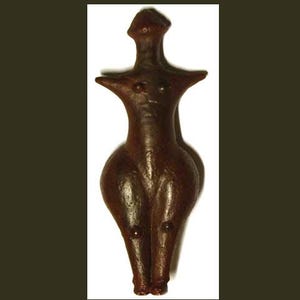 May include: A brown, stylized figurine of a female form. The figure has a rounded body, small arms, and a head with a pointed top. The figurine is made of a dark brown material and has a smooth, polished appearance.
