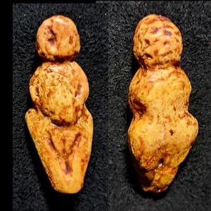 May include: Two small, tan-colored sculptures of a human figure. The figures have rounded shapes, with a head, torso, and legs. The sculptures are on a dark background. One figure is viewed from the front, the other from the side.
