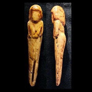 May include: Two small, carved figurines of a human form. The figures are a light tan color with a smooth texture, and appear to be ancient artifacts. One figure is viewed from the front, the other from the side.