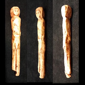 May include: Three views of a small, carved figurine. The figure is a light beige color with darker brown accents. It appears to be a stylized human form, with a simple face and elongated body. The figure is standing, with arms at its sides.