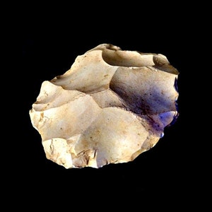 May include: A light brown, irregularly shaped stone tool with a rough, textured surface. The tool has been chipped and shaped, likely for use as a hand axe or scraper.